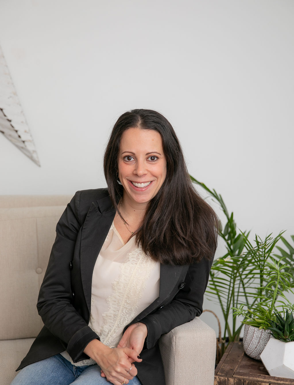 Nikki has brown hair and brown eyes and is sitting on a couch, wearing a blazer and jeans