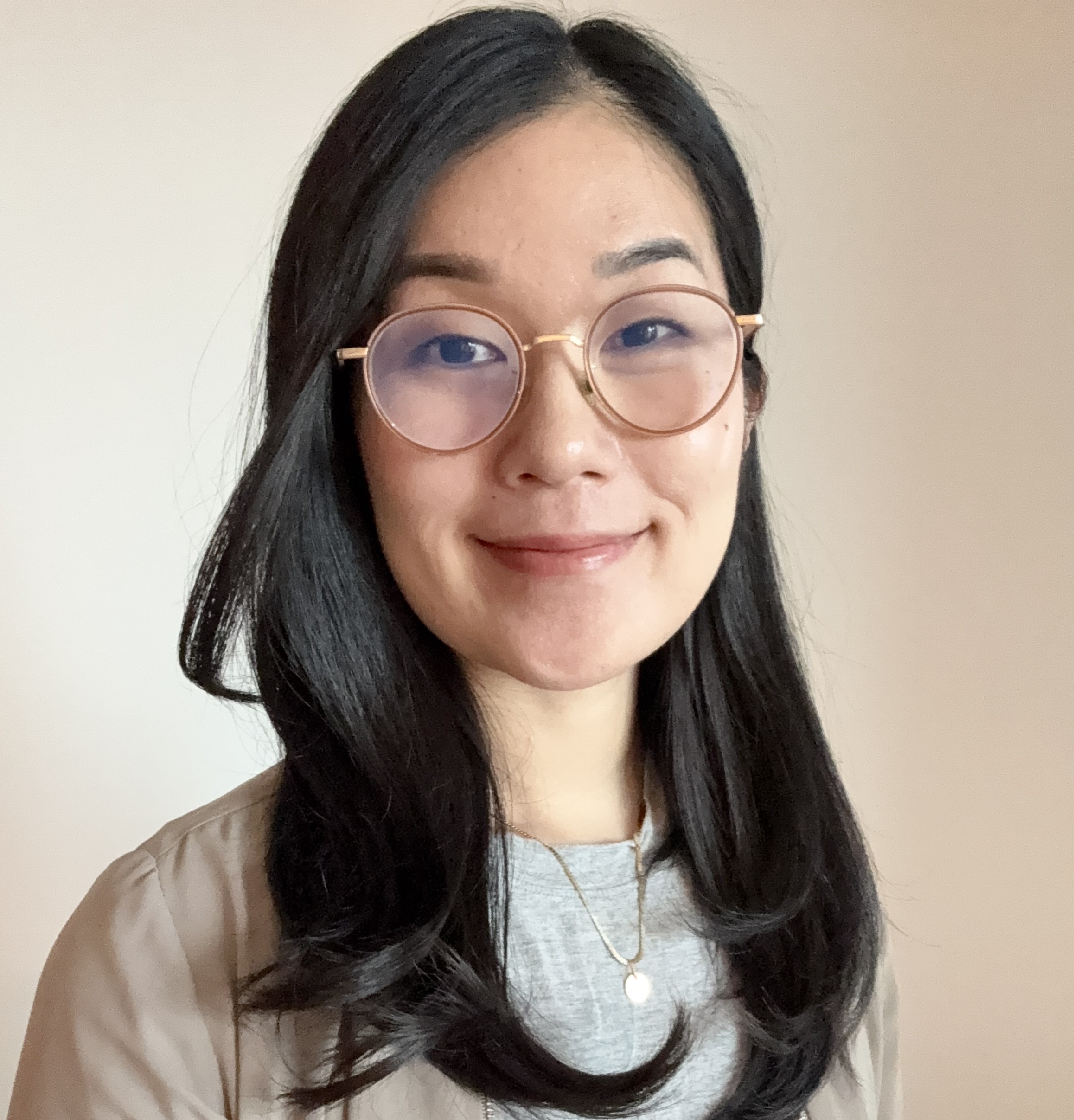 An East-Asian woman, medium length dark brown hair, wearing circular rimmed glasses, smiling, in front of a white wall.