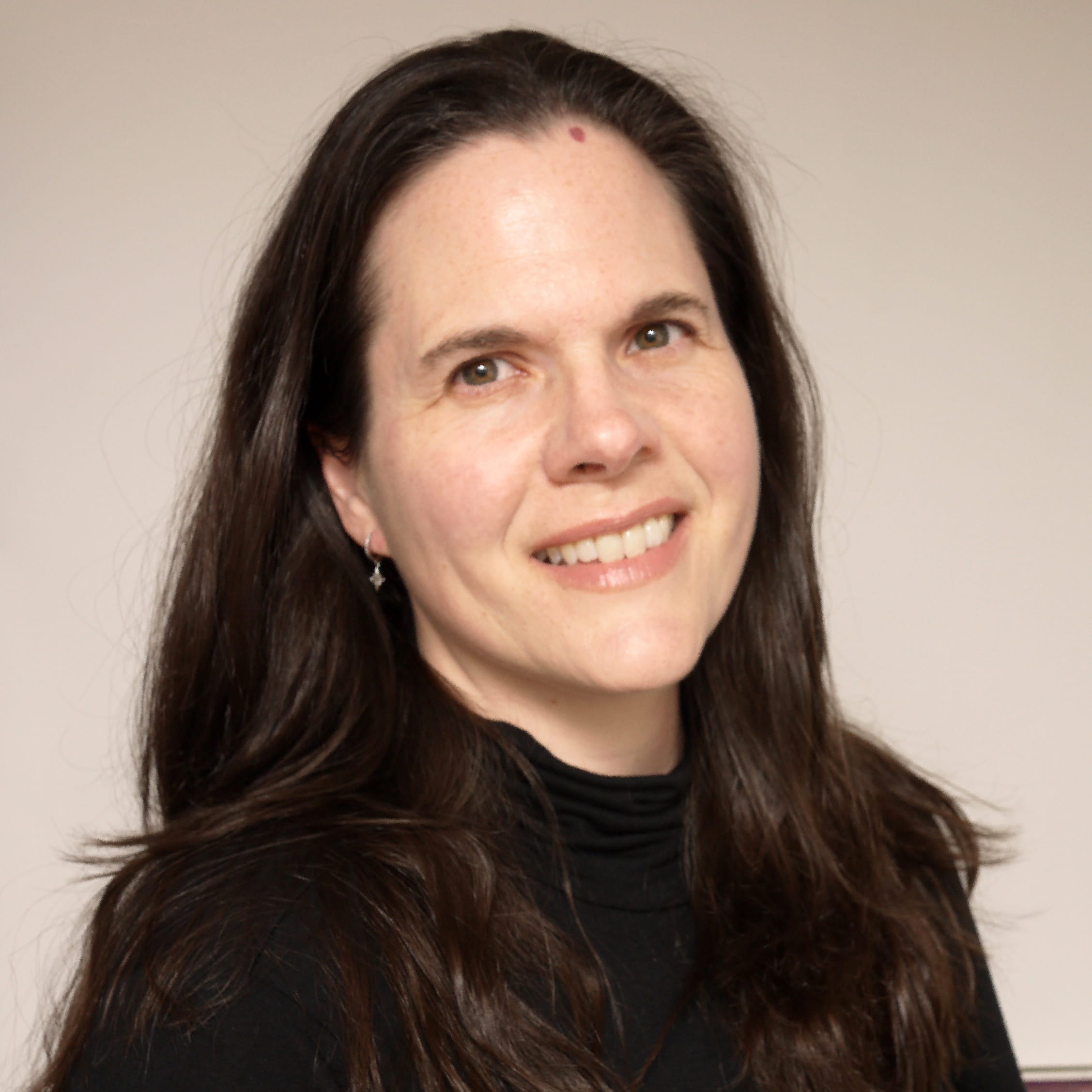 A woman with long dark hair and a slight smile poses against a light background, wearing a black turtleneck and silver earrings. Her expression is neutral and confident.